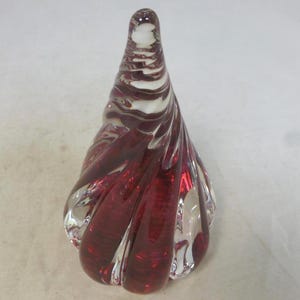 Peut inclure: Un objet décoratif en verre en forme de goutte d'eau tourbillonnante. Le verre est d'un rouge profond, avec des sections transparentes qui laissent passer la lumière. L'objet a une finition lisse et polie.