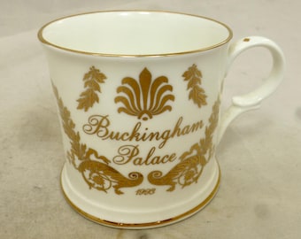 Puede incluir: Taza de cerámica blanca con borde dorado y diseño dorado. El diseño presenta un motivo floral estilizado y el texto "Buckingham Palace 1968".