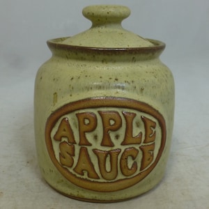 May include: A ceramic jar with a lid, decorated with a brown oval and the words "APPLE SAUCE" in a retro font. The jar is a light green color with brown speckles.