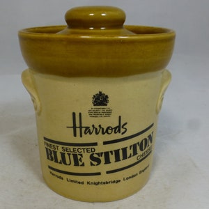 May include: A ceramic jar with a lid, featuring the Harrods logo and the text "Finest Selected Blue Stilton Cheese". The jar is beige with a brown lid and has two handles on the sides.