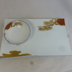 Puede incluir: Un plato de servir de porcelana blanca con un tazón pequeño adjunto. El plato presenta un diseño dorado y rojo de una rama de pino con una forma de nube estilizada en la parte inferior.