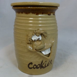 Peut inclure: Une jarre à biscuits en céramique avec un glaçage brun et un visage fantaisiste. La jarre a un couvercle et dit "Cookies" sur le devant.