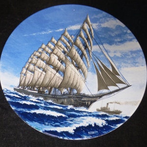 Könnte beinhalten: Ein antiker Keramikteller mit einem Gemälde eines großen Segelschiffs mit weißen Segeln vor blauem Himmel und Meer. Das Schiff segelt auf einem blauen Meer mit weißen Wellen. Der Teller hat einen blauen Rand und einen weißen Hintergrund.