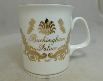 Puede incluir: Taza de cerámica blanca con un diseño dorado con las palabras "Buckingham Palace" y el año 1993. La taza tiene un asa curva y un borde dorado. El diseño incluye elementos florales y ornamentales.