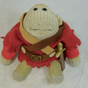 May include: A knitted plush toy in the shape of a creature with a beige head and limbs, and a red tunic. It has a brown belt with a gold-colored buckle and a small sword. The toy has black eyes.