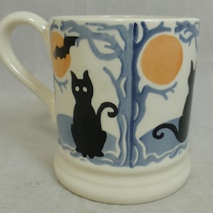 May include: Cream-colored ceramic mug with a handle, featuring a Halloween design. The mug has black cat silhouettes, orange moons, and blue tree and water details. The mug is a cylindrical shape.