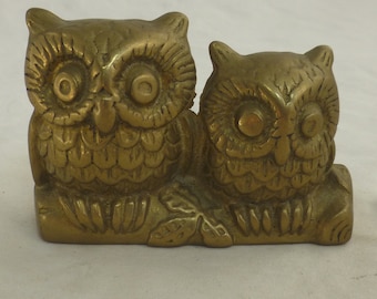 May include: A brass sculpture featuring two owls perched together. The owls have detailed feather textures and large, round eyes. The sculpture is a warm gold color and sits on a small base with leaf details.