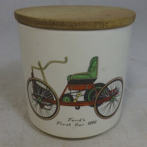 Könnte beinhalten: Weißer Keramiktopf mit Holzdeckel. Der Topf zeigt eine farbenfrohe Illustration von Fords erstem Auto von 1896. Das Auto ist mit grünen, roten und gelben Akzenten dargestellt. Ein dekorativer Artikel für das Zuhause.