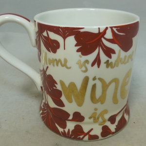 Op de afbeelding: Witte keramische mok met een rood en gouden ontwerp. De mok heeft een handvat en zegt "Home is where wine is" in gouden letters.