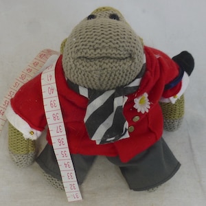 May include: A knitted monkey wearing a red jacket, a black and white striped scarf, and grey trousers. The monkey has a white flower pinned to its jacket. A measuring tape is wrapped around the monkey's arm.