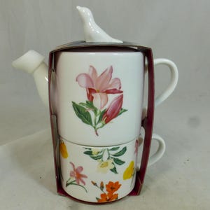 Puede incluir: Juego de té de cerámica blanca con una tetera y una taza. La tetera tiene una tapa con forma de pájaro y un diseño floral con flores rosas. La taza tiene un estampado floral con flores amarillas y naranjas.