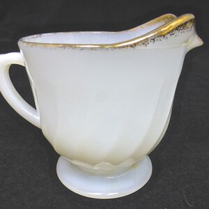 Peut inclure: Une crémier en verre de lait blanc avec un bord doré et une anse. La crémier a un design cannelé et une base arrondie.