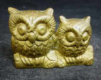 May include: Two gold-colored owl figurines perched on a branch. The owls have detailed features, including large eyes and feathery wings.