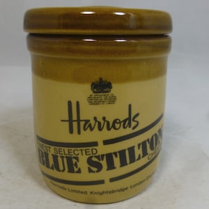 May include: A brown ceramic jar with a lid. The jar has a white label with black text that reads "Harrods Finest Selected Blue Stilton Cheese Harrods Limited Knightsbridge London England".