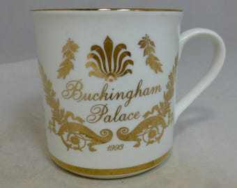 Puede incluir: Taza de cerámica blanca con ribete dorado y un diseño dorado con las palabras "Buckingham Palace" y el año 1998. La taza tiene un asa curva y está decorada con detalles florales y ornamentales.