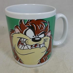 Może przedstawiać: Biały ceramiczny kubek z postacią z kreskówki Tasmanian Devil. Kubek ma zielone tło z twarzą postaci w kolorze brązowym, beżowym i białym. Kubek ma biały uchwyt i białe wnętrze.