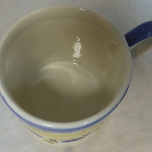 May include: A white ceramic mug with a blue rim and handle. The mug has a floral design on the bottom.