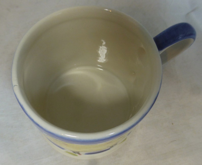 May include: A white ceramic mug with a blue rim and handle. The mug has a floral design on the bottom.