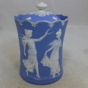 May include: A blue and white ceramic jar with a lid. The jar features a raised design of two women in white, one holding a branch with leaves. The jar has a scalloped rim.