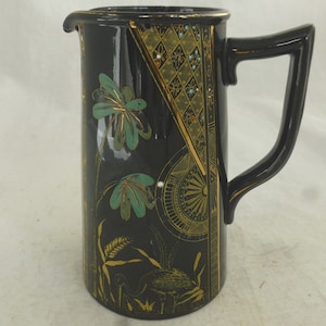 Puede incluir: Jarra de cerámica negra con un diseño floral dorado y verde. La jarra tiene una boca ancha y un asa.