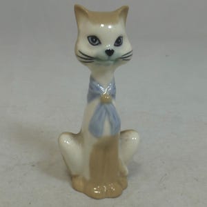 May include: A ceramic cat figurine with a light brown and white color scheme. The cat has blue eyes, black whiskers, and a light blue bow around its neck. The figurine is in a seated position.
