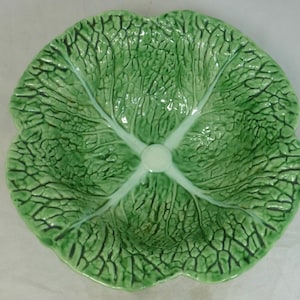 May include: A round ceramic bowl shaped like a green cabbage leaf. The surface features a glossy finish with intricate, raised vein patterns that mimic the texture of a real vegetable leaf, radiating from a smooth, circular centre point.