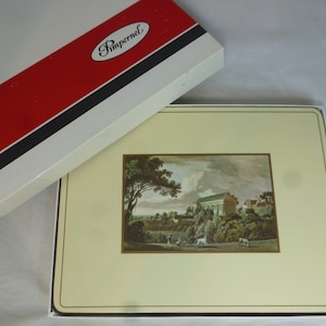 Könnte beinhalten: Ein Set mit vier beigefarbenen Platzsets mit goldfarbenem Rand. Jedes Platzset zeigt eine Vintage-Illustration eines Gebäudes mit einem grünen Baum und einem bewölkten Himmel. Die Platzsets sind in einer weißen Schachtel mit einem roten und schwarzen Streifen verpackt, auf der das Wort "Pimpernel" in Schwarz gedruckt ist.