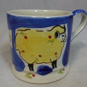 May include: A white ceramic mug with a blue and yellow hand-painted design of a sheep. The sheep is yellow with red spots and black legs. The mug has a white handle.