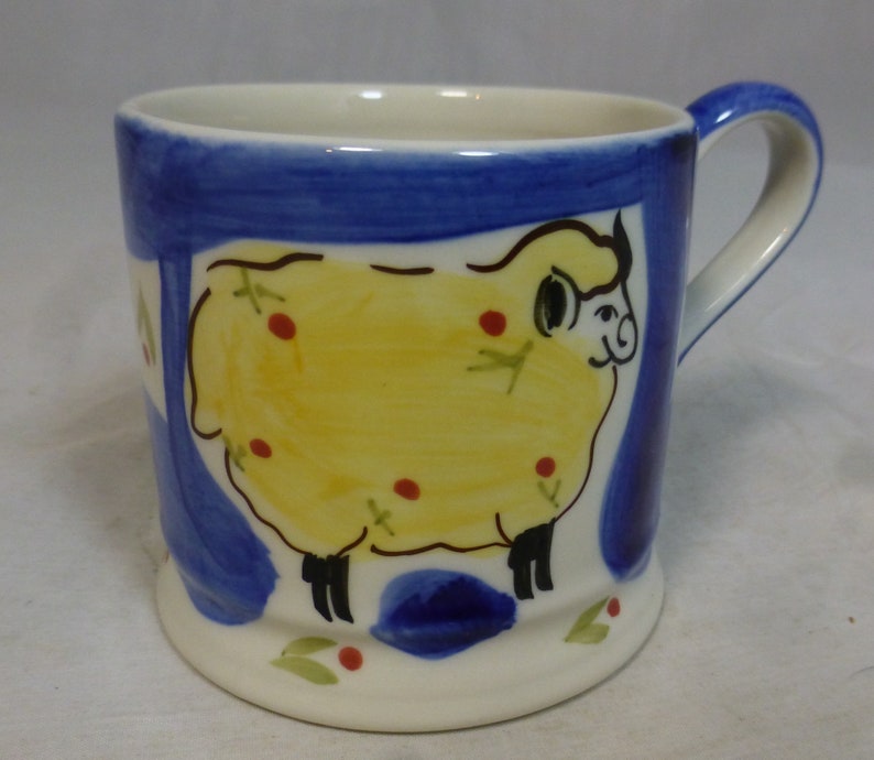 May include: A white ceramic mug with a blue and yellow hand-painted design of a sheep. The sheep is yellow with red spots and black legs. The mug has a white handle.