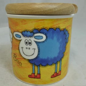 May include: A ceramic jar with a wooden lid features a cartoon blue sheep with a white face and red shoes. The jar has a yellow background with the word "baa" written on it.