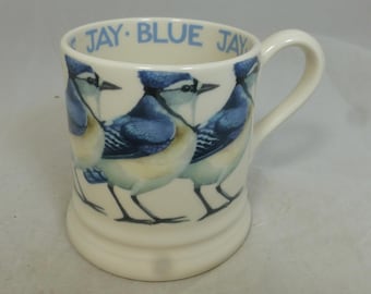 Può includere: Tazza in ceramica color crema con illustrazioni di ghiandaie blu. La tazza ha un manico curvo e le parole "JAY BLUE JAY" in blu attorno al bordo. Le ghiandaie blu sono raffigurate con dettagli blu, bianchi e neri.