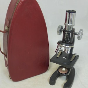 May include: A vintage black and silver microscope with a red carrying case. The microscope has adjustable knobs and a stage for slides. The case is a deep red with a handle and zip.