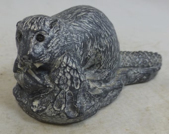 May include: A gray stone sculpture of a beaver sitting on a log, holding a branch in its mouth. The beaver's fur is detailed, and its eyes are black.