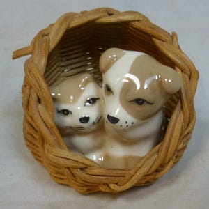 May include: A ceramic figurine of a puppy and kitten nestled together in a small woven basket. The puppy and kitten are tan and white with black facial features. The basket is made of woven, light brown wicker.