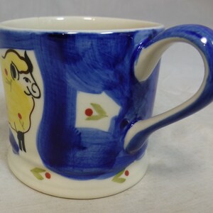 May include: A white ceramic mug with a blue and yellow sheep design. The mug has a white handle and a blue rim.