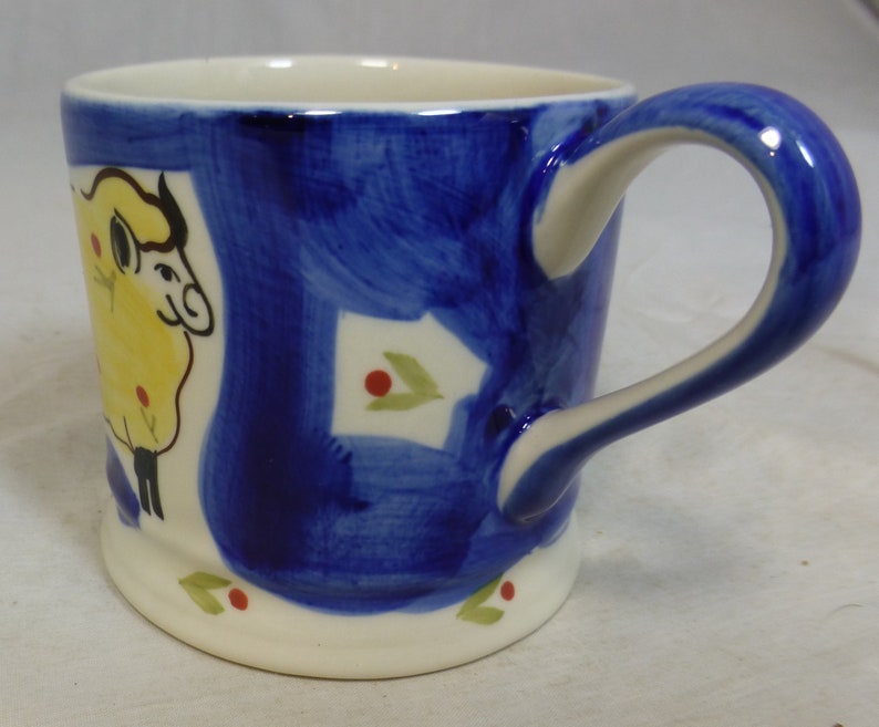 May include: A white ceramic mug with a blue and yellow sheep design. The mug has a white handle and a blue rim.