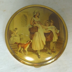 Op de afbeelding: Een rond, goudkleurig compact met een geschilderd tafereel van vier mensen en een hond. De scène toont een vrouw in een witte jurk en een hoed, twee kinderen en een hond voor een gebouw.