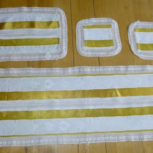 Puede incluir: Un juego de mantelería blanca con ribete de cinta de satén dorado y borde de encaje. El juego incluye un camino de mesa y dos cuadrados más pequeños.