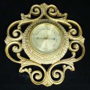 May include: A vintage gold-colored barometer with an ornate, scrollwork frame. The circular gauge features a brass face with a needle indicating atmospheric pressure. The dial reads "Falls for Storms or Rain" and "Rises for Fair or Dry."