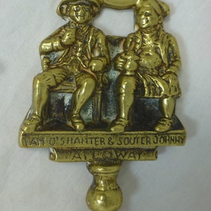 May include: Brass doorstop featuring two figures sitting on a bench. The figures are labeled "Tam O'Shanter & Souter Johnny" and "Alloway".