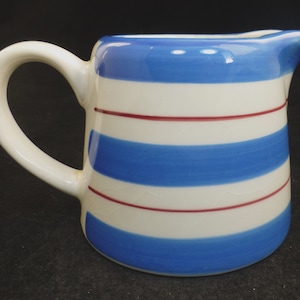 以下が含まれることがあります： 青と赤のストライプ模様の白いセラミック製のクリーマー。クリーマーにはハンドルと注ぎ口があります。