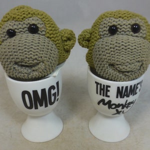 Puede incluir: Dos soportes para huevos de cabeza de mono tejidos. Una taza dice "OMG!" y la otra dice "The Name's Monkey".