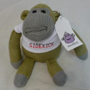 Puede incluir: Un peluche de mono tejido en verde oliva y gris, con una camiseta blanca con la palabra "SIDEKICK" en letras rojas y negras. Tiene una etiqueta con la cara de un mono morado y la palabra "monkey".