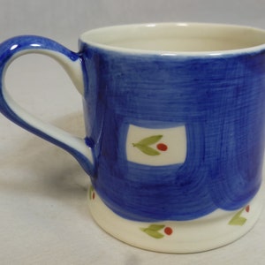 May include: A blue ceramic mug with a white handle and a white rim. The mug has a painted design of white squares with green leaves and red berries.