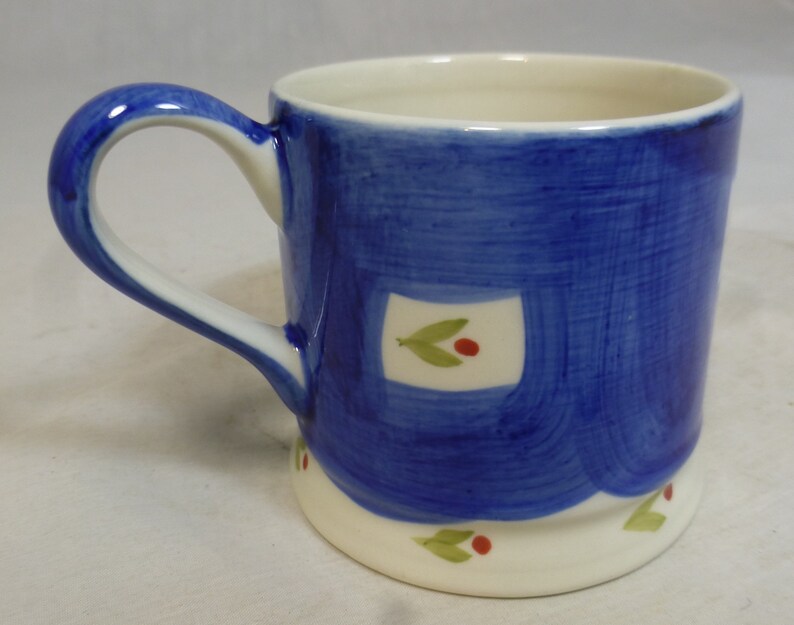 May include: A blue ceramic mug with a white handle and a white rim. The mug has a painted design of white squares with green leaves and red berries.