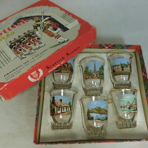 Puede incluir: Un juego de seis vasos de chupito de vidrio transparente con bordes dorados. Cada vaso presenta una imagen diferente de una escena escocesa, incluyendo un castillo, una iglesia, un puente y una cabaña. Los vasos están empaquetados en una caja roja y blanca con las palabras "Scottish Scenes Drinking Set" impresas en ella.