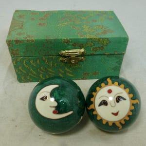 May include: Two dark green spherical meditation balls featuring a cream-coloured crescent moon on one and a sun face on the other, accompanied by a rectangular green fabric-covered storage box with a gold-toned metal latch closure.