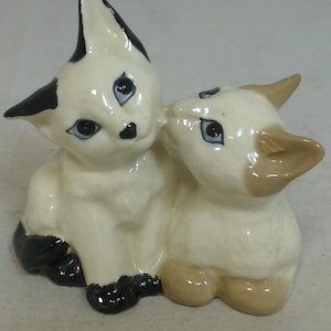May include: A ceramic figurine of two stylized cats. One cat is white with black ears and paws, the other is cream-colored with tan ears and paws. Both have blue eyes and are posed together.