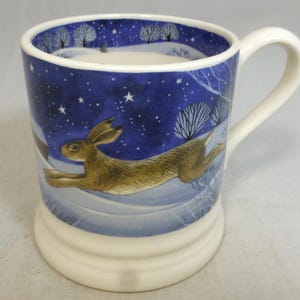 May include: A ceramic mug with a winter scene featuring a running hare. The mug has a white handle and base, with a blue background depicting a starry night and snow-covered ground. The hare is brown and tan.