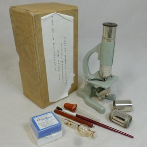 May include: A light gray metal microscope with a matching storage box labeled Philip Harris Limited D.I.Y. Microscope and Slide Kit. The set includes two silver metal cylinders, a glass dropper with a red top, a glass pipette, and a red stirring rod.